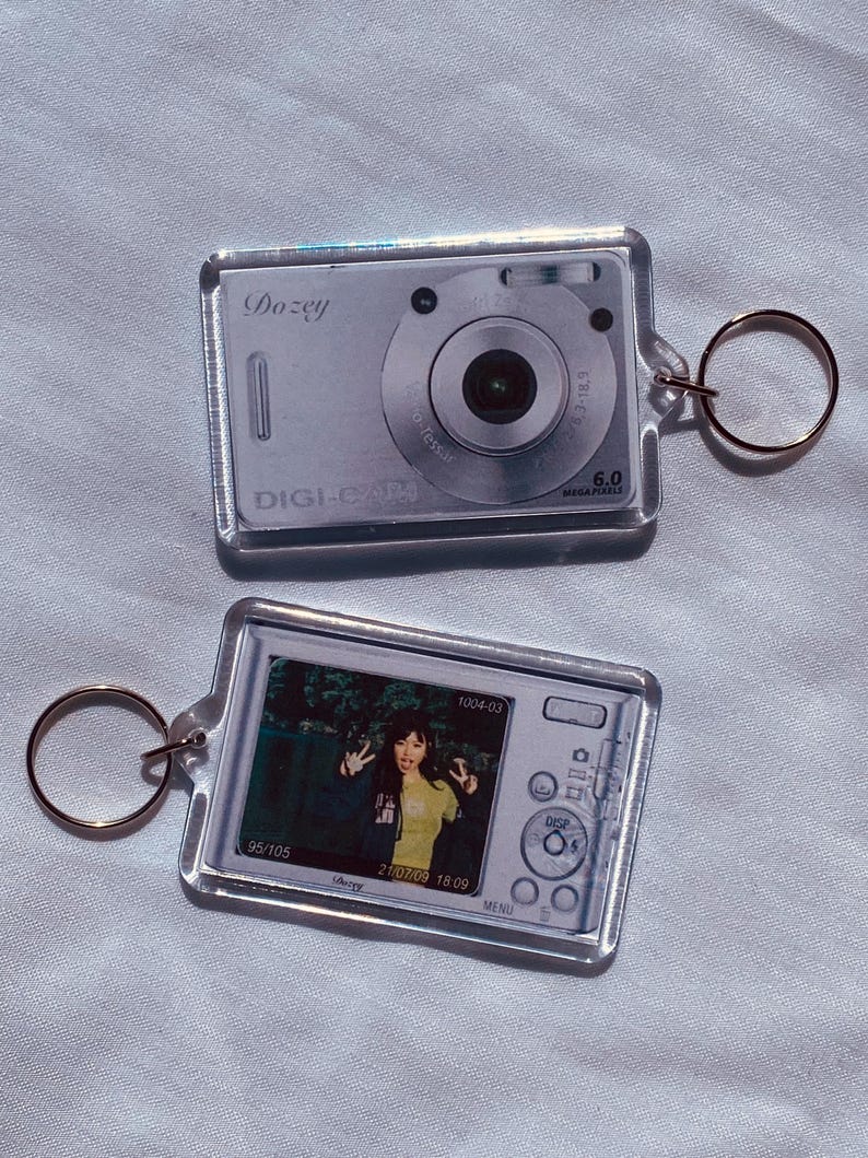 May include: Two clear acrylic keychains shaped like digital cameras. One keychain shows a camera with the text "Dozey" and "DIGI-CAM". The other keychain has a photo of a person. Both keychains have a gold-colored ring.