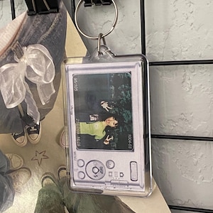 May include: Clear rectangular keychain with a photo of a person making a peace sign. The keychain has a silver camera design with the text "95/105", "21/07/09 18:09", and "1004-03". The keychain is attached to a silver keyring.