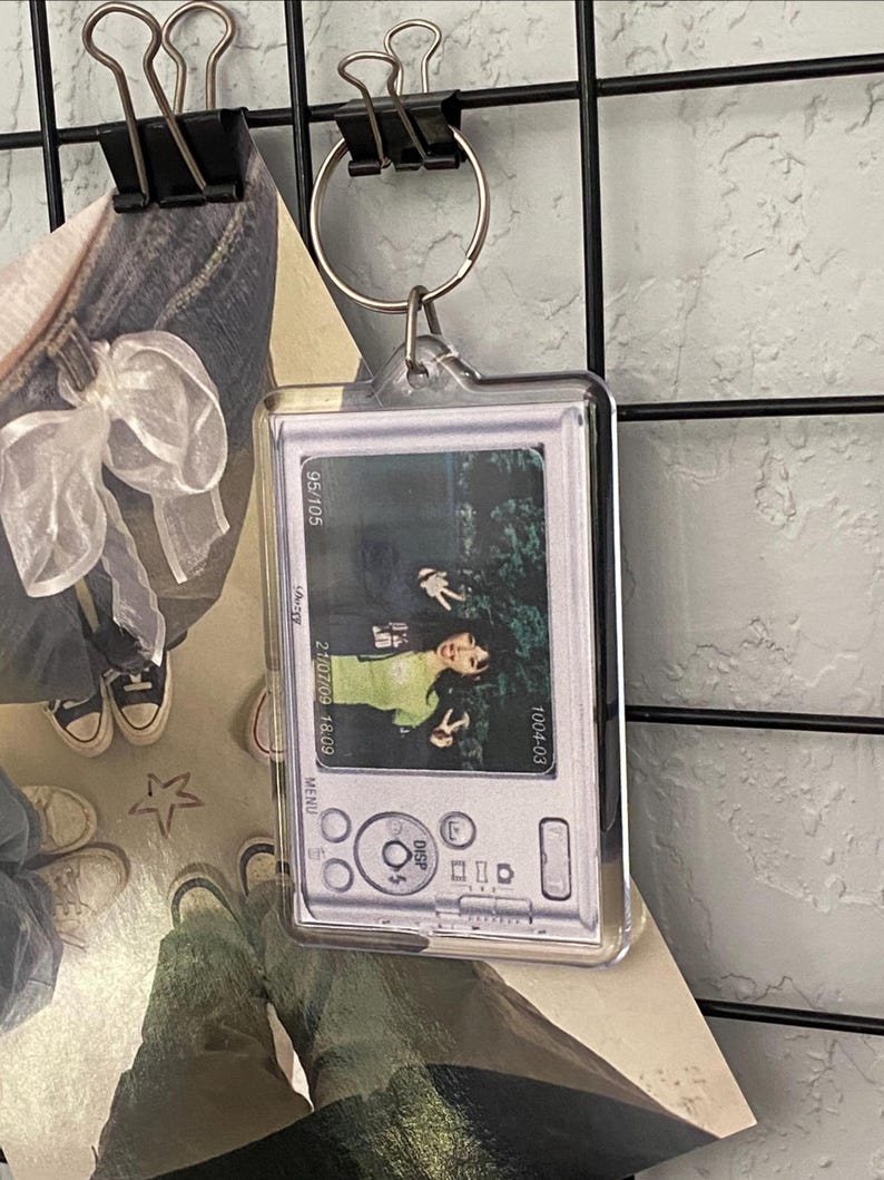 May include: Clear rectangular keychain with a photo of a person making a peace sign. The keychain has a silver camera design with the text "95/105", "21/07/09 18:09", and "1004-03". The keychain is attached to a silver keyring.