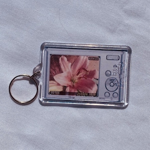 May include: A clear rectangular keychain with a gold-colored ring. The keychain features a small image of a digital camera displaying a pink flower. The camera screen shows the numbers "1004-03", "95/105", and "21/07/09 18:09".