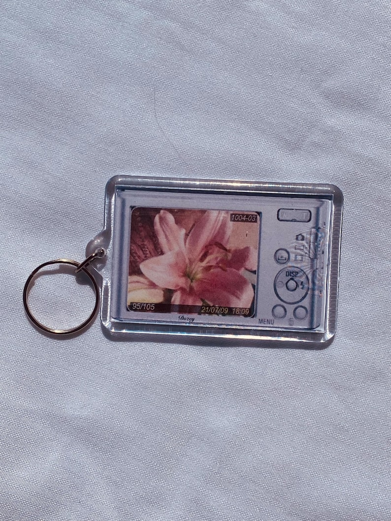 May include: A clear rectangular keychain with a gold-colored ring. The keychain features a small image of a digital camera displaying a pink flower. The camera screen shows the numbers "1004-03", "95/105", and "21/07/09 18:09".