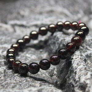 May include: A dark red gemstone bracelet made of round beads. The bracelet is on a gray textured surface.