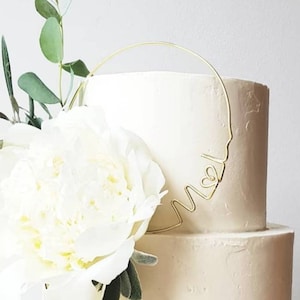 Könnte beinhalten: Eine zweistöckige weiße Hochzeitstorte mit einem goldenen Draht-Topper in Form eines Herzens und den Buchstaben "M & W". Eine weiße Pfingstrose und Grün sind an der Seite der Torte.