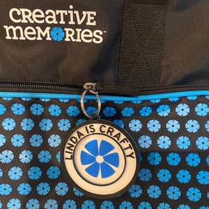 May include: Black zippered tote bag with a blue trim and a blue and black floral pattern. The bag has a silver zipper pull with a round black tag that says "Linda is Crafty" with a blue flower in the center.