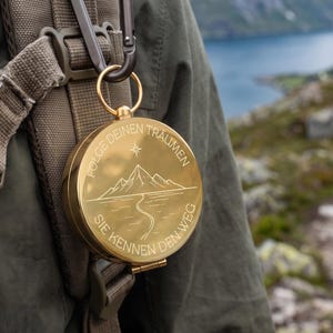 May include: A round gold-coloured metal compass attached to a dark olive green backpack strap with a black carabiner. The compass lid features an engraved mountain landscape, a winding path, and the German text: FOLGE DEINEN TRÄUMEN SIE KENNEN DEN WEG.