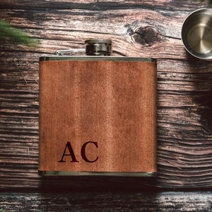 May include: A rectangular metal flask wrapped in a smooth, medium-brown wood veneer finish. The front surface features the engraved capital letters AC in a dark, serif font. The flask has a silver-toned metal screw-top cap at the centre top.