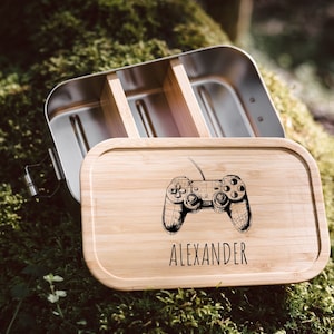 Op de afbeelding: Een lunchbox van roestvrij staal met een bamboe deksel. Het deksel heeft een zwart-witte illustratie van een videogamecontroller en de naam "Alexander" gegraveerd.