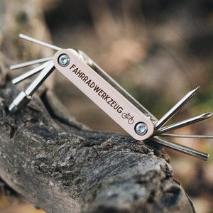 Op de afbeelding: Een multitool voor fietsen met een houten handvat en de tekst "FAHRRADWERKZEUG" erop gegraveerd. De tool heeft verschillende hulpstukken, waaronder een spaakmoersleutel, een kettinggereedschap en een bandenlichter.