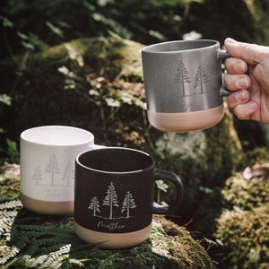 Puede incluir: Tres tazas de cerámica con un diseño de bosque. Una es sostenida por una persona. Las tazas son grises, negras y blancas con un acabado moteado y una base beige. Cada taza tiene un gráfico de árbol y un nombre.