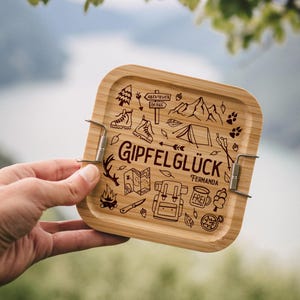 Könnte beinhalten: Eine quadratische, hellbraune Bambus-Lunchbox mit Metallverschlüssen. Der Deckel ist mit Camping-Illustrationen und dem deutschen Text "Gipfelglück" und "Fernanda" graviert. Das Design umfasst ein Zelt, Berge und Wanderschuhe.