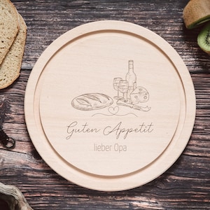 Puede incluir: Una tabla de cortar redonda de madera con un diseño grabado de pan, queso, vino y vasos. El texto "Guten Appetit lieber Opa" está grabado en la tabla.