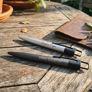 Può includere: Due penne con fusti neri e argentati sono su una superficie di legno. La penna argentata ha il testo "Bester Papa" e la penna nera "HANNAS GLÜCKSSTIFT". Un portafoglio in pelle marrone è sullo sfondo.