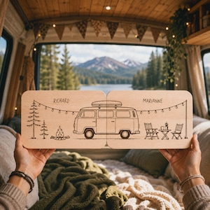 Könnte beinhalten: Ein rechteckiges Holzschild mit einer lasergravierten Illustration eines Campingbusses, Tannenbäumen, einem Lagerfeuer und Gartenmöbeln. Die Namen Richard und Marianne sind über dem Bus eingraviert, ergänzt durch eine Lichterkette.