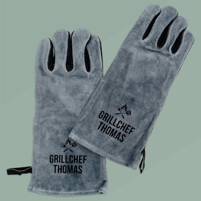 Op de afbeelding: Een paar grijze su&egrave;de barbecuehandschoenen met zwarte accenten. Op elke handschoen staat in het zwart de tekst "GRILLCHEF THOMAS", samen met een afbeelding van een spatel en vlammen. De handschoenen beschermen de handen tijdens het grillen.