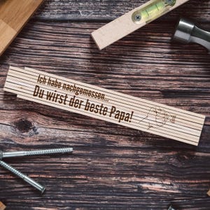 Könnte beinhalten: Ein Holzlineal mit dem Text "Ich habe nachgemessen... Du wirst der beste Papa!" Das Lineal liegt auf einer braunen Holzoberfläche mit einem Hammer, Schrauben und einer Wasserwaage.