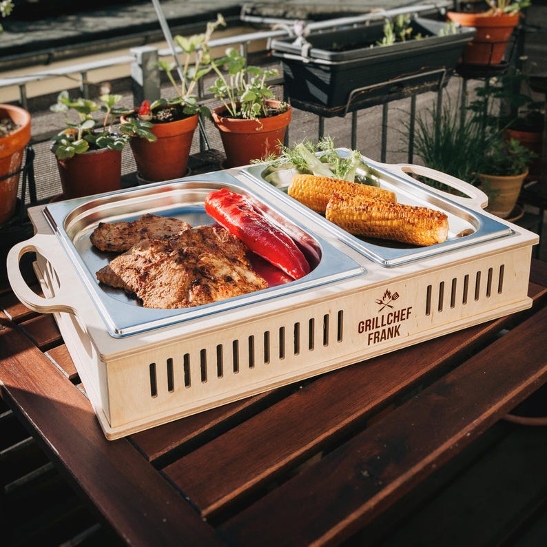 May include: A wooden grilling tray with two stainless steel compartments, one with grilled meat and red peppers, the other with corn on the cob and greens. The tray has handles and the text "GRILLCHEF FRANK".