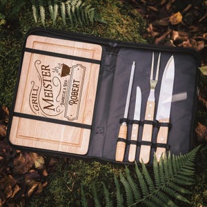 Può includere: Un set da barbecue in legno con tagliere e utensili. Il tagliere presenta le incisioni "Grill Meister" e "Robert". Il set include un coltello, una forchetta e un affilatore, tutti con manici in legno, all'interno di una custodia nera.