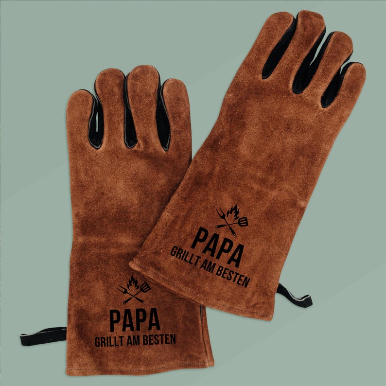 May include: Brown leather grilling gloves with black accents. The gloves have the text "PAPA GRILLT AM BESTEN" printed in black, along with a graphic of a grill and utensils. The gloves are designed to protect hands from heat.
