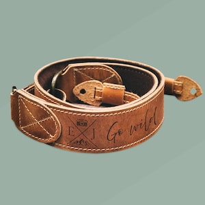 Può includere: Tracolla per fotocamera in pelle marrone con le iniziali "E.J." e un'icona della fotocamera, insieme alle parole "Go wild" incise su di essa.