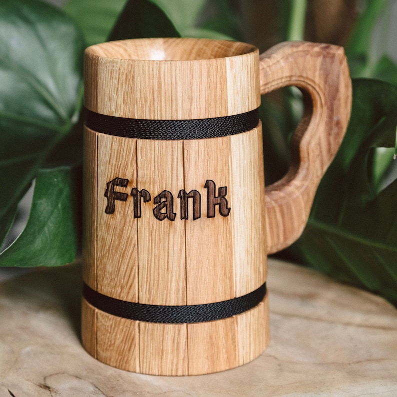 Op de afbeelding: Houten bierpul met de naam "Frank" gegraveerd op de voorkant. De pul is gemaakt van lichtgekleurd hout met een grote handgreep en is omcirkeld door twee zwarte banden. Ontworpen om dranken in te bewaren.