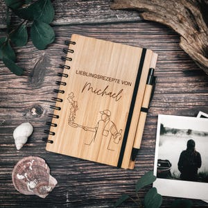 Könnte beinhalten: Ein spiralgebundenes Notizbuch mit einem hellbraunen Bambuseinband, verziert mit einer schwarzen Strichzeichnung einer Person, die mit einer Pfanne kocht, sowie dem Text LIEBLINGSREZEPTE VON Michael. Ein passender Bambuskugelschreiber steckt in einer schwarzen Schlaufe.