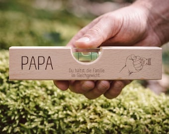 May include: A wooden level with the word "PAPA" engraved on it. The level has a bubble vial and a drawing of a hand holding a baby's hand. The text "Du hältst die Familie im Gleichgewicht" is also engraved on the level.