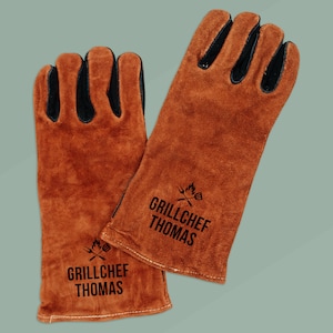 Op de afbeelding: Een paar bruin lederen barbecuehandschoenen met zwarte accenten op de vingers. De handschoenen hebben de tekst "GRILLCHEF THOMAS" in het zwart gedrukt, samen met een afbeelding van een grill en keukengerei.