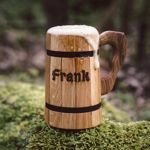 Op de afbeelding: Een houten bierpul met de naam "Frank" in de voorkant gegraveerd. De pul is gevuld met bier en schuim stroomt over. De handgreep is van hout. De pul staat op groen mos.