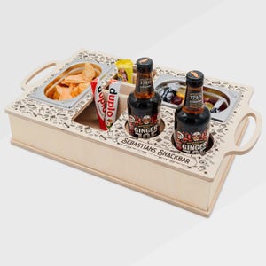May include: A light-colored wooden snack tray with cutouts for drinks and snacks. The tray has two metal containers filled with chips and fruit. Two bottles of ginger beer and a few snack packets are also visible. The tray has handles and the text "SEBASTIANS SNACKBAR".