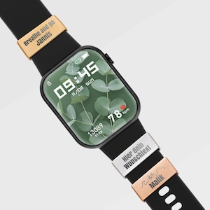 Op de afbeelding: Een zwarte smartwatch met een groen display dat de tijd, datum en stappen weergeeft. De horlogeband heeft gouden en zilveren accenten met de tekst "Breathe and go Jannis" en "Hier dein Wunschtext!".