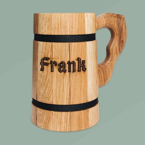 Op de afbeelding: Een houten bierpul met de naam "Frank" in de voorkant gegraveerd. De pul is gemaakt van lichtgekleurd hout met donkere banden en een gebogen handvat.
