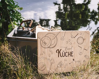 May include: A wooden sign with the words "Bord Küche" and kitchen-themed illustrations, including plates, utensils, and a coffee maker. A gray storage bin filled with kitchenware sits behind the sign, near a body of water.