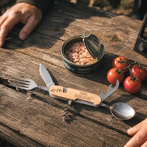 Op de afbeelding: Een multifunctioneel kampeerbestek met een houten handvat gegraveerd met bergpictogrammen en de tekst "Franks KLEINE RETTUNG", voorzien van een uitklapbare vork, mes, lepel, kurkentrekker en flesopener, naast een blik tonijn en vier tomaten.