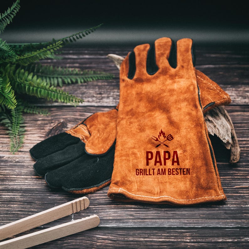 May include: A pair of brown leather grilling gloves with black suede palms. The gloves have the text "PAPA GRILLT AM BESTEN" printed in red, along with a graphic of a flame and crossed grill tools. Wooden tongs are in the foreground.