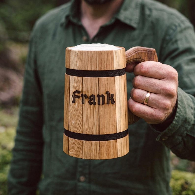 Op de afbeelding: Houten bierpul met de naam "Frank" gegraveerd op de voorkant. De pul is gemaakt van lichtgekleurd hout met donkere banden en een handvat. De pul is gevuld met bier en schuim.