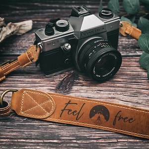 Può includere: Un cinturino per fotocamera in pelle marrone con il testo "feel free" e una scena di montagna con un falò e stelle. Il cinturino è attaccato a una fotocamera Praktica MTL 5 B argentata e nera.