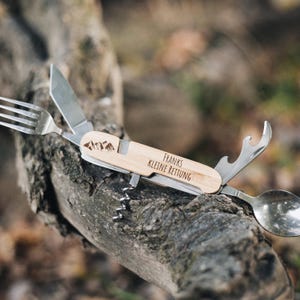 Könnte beinhalten: Ein Multifunktions-Camping-Besteck mit einem Holzgriff, der mit "Franks Kleine Rettung" und einem Bergpanorama-Design graviert ist. Das Werkzeug beinhaltet eine Gabel, einen Löffel, ein Messer, einen Flaschenöffner und einen Korkenzieher.