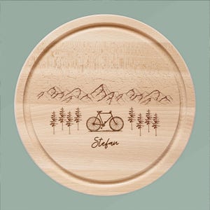 May include: A round wooden cutting board with a mountain and bicycle design. The board has a light wood color and features an engraved mountain range, trees, and a bicycle. The name "Stefan" is engraved below the bicycle.