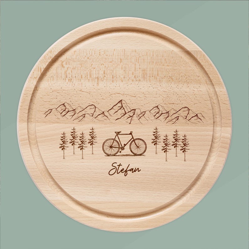 May include: A round wooden cutting board with a mountain and bicycle design. The board has a light wood color and features an engraved mountain range, trees, and a bicycle. The name "Stefan" is engraved below the bicycle.