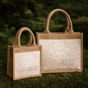 Puede incluir: Dos bolsas de mano de yute con asas. Las bolsas son de color beige natural con un panel rectangular blanco en la parte delantera. El panel presenta un dise&ntilde;o de flor de loto blanca y el texto "Wellness Auszeit" en una fuente cursiva.