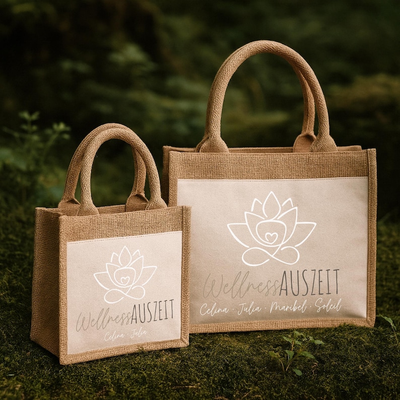 Puede incluir: Dos bolsas de mano de yute con asas. Las bolsas son de color beige natural con un panel rectangular blanco en la parte delantera. El panel presenta un dise&ntilde;o de flor de loto blanca y el texto "Wellness Auszeit" en una fuente cursiva.