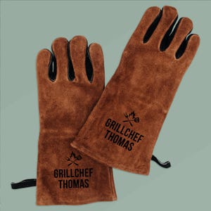 Op de afbeelding: Een paar bruine leren barbecuehandschoenen met zwarte accenten op de vingers. De handschoenen hebben de tekst "GRILLCHEF THOMAS" in het zwart gedrukt, samen met een afbeelding van een grill en keukengerei. De handschoenen beschermen de handen tegen hitte.