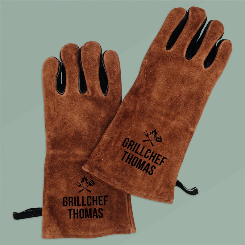 Op de afbeelding: Een paar bruine leren barbecuehandschoenen met zwarte accenten op de vingers. De handschoenen hebben de tekst "GRILLCHEF THOMAS" in het zwart gedrukt, samen met een afbeelding van een grill en keukengerei. De handschoenen beschermen de handen tegen hitte.
