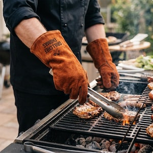 Op de afbeelding: Bruine leren grillhandschoenen met de tekst "GRILLCHEF THOMAS" in het zwart. De persoon gebruikt een tang om voedsel op een houtskoolgrill te hanteren. De grill is gevuld met steaks en worstjes.