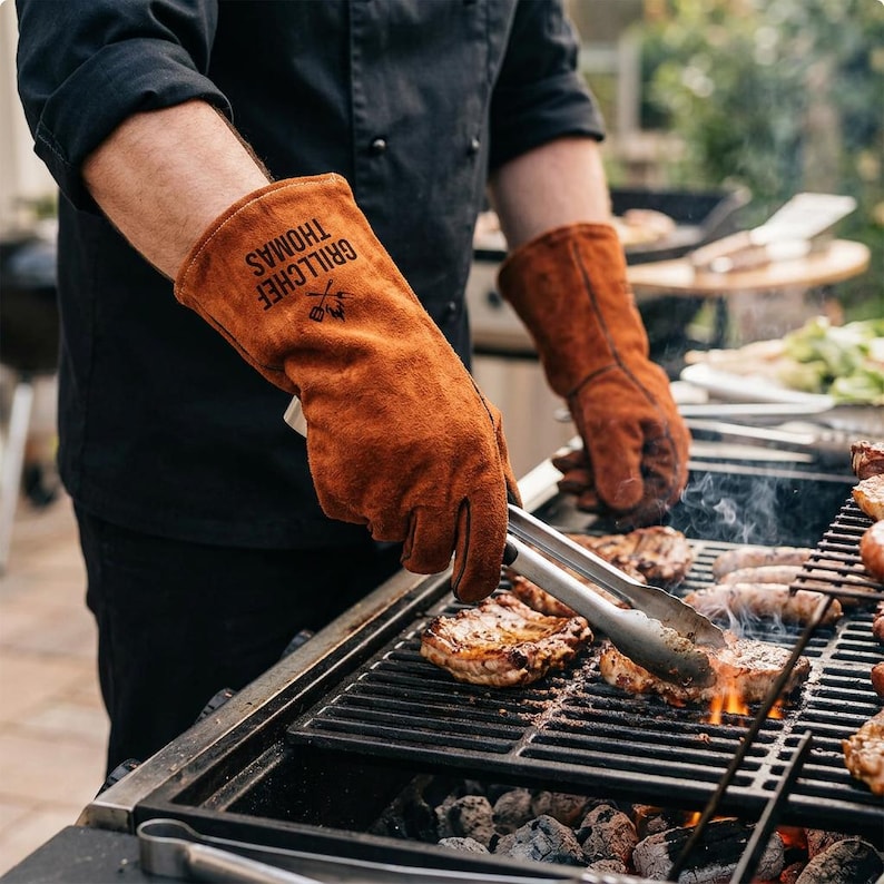 Op de afbeelding: Bruine leren grillhandschoenen met de tekst "GRILLCHEF THOMAS" in het zwart. De persoon gebruikt een tang om voedsel op een houtskoolgrill te hanteren. De grill is gevuld met steaks en worstjes.