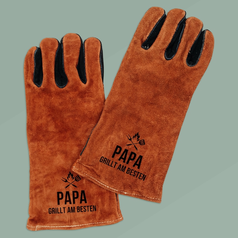 May include: A pair of brown leather grilling gloves with black accents on the fingers. The gloves have the text "PAPA GRILLT AM BESTEN" printed in black, along with a graphic of crossed grilling tools.