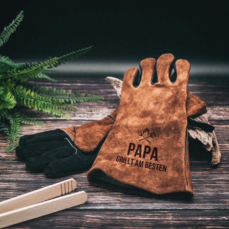 May include: Brown leather grilling gloves with black accents and the text "PAPA GRILLT AM BESTEN". The gloves are resting on a wooden surface with a pair of wooden tongs.