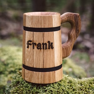 Op de afbeelding: Een houten bierpul met de naam "Frank" in de voorkant gegraveerd. De pul is gemaakt van lichtbruin hout met een donkere handgreep en twee zwarte banden. De pul staat op een bedje van groen mos.