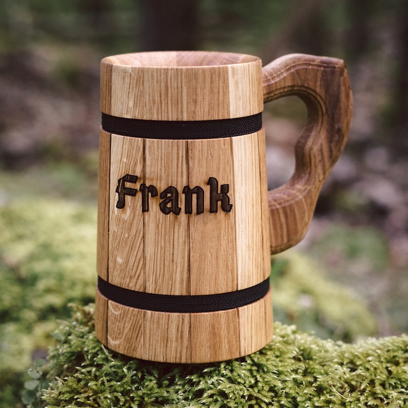 Op de afbeelding: Een houten bierpul met de naam "Frank" in de voorkant gegraveerd. De pul is gemaakt van lichtbruin hout met een donkere handgreep en twee zwarte banden. De pul staat op een bedje van groen mos.