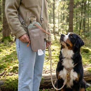 Könnte beinhalten: Eine beige Umhängetasche mit einer Wasserflasche und einem Spender für Hundekotbeutel. Auf der Tasche steht der Name "BALOU". Ein Hund schaut die Person an, die die Leine hält. Der Hund ist schwarz, braun und weiß.